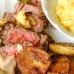 A white plate with grilled steak and potatoes, with a dish of creamed corn on the side.