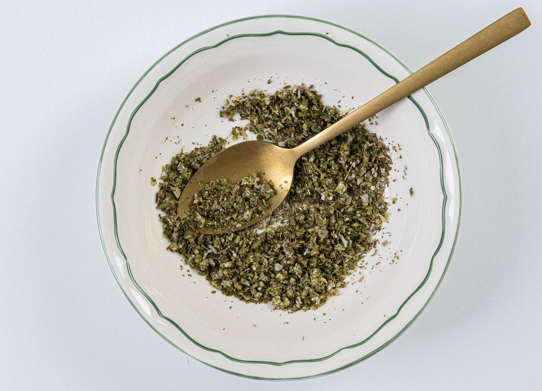 A white bowl with a green rim holds a mix of dried herbs, with a gold spoon resting inside.
