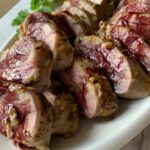 Sliced pork tenderloin with grainy mustard glaze and red wine sauce on a white plate, garnished with fresh herbs.