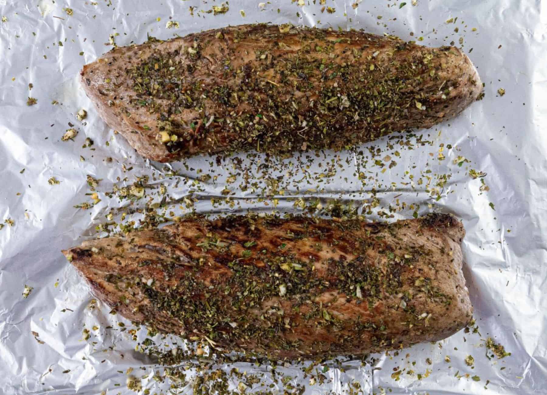 Two pork tenderloins seasoned with herbs and spices rest on aluminum foil.