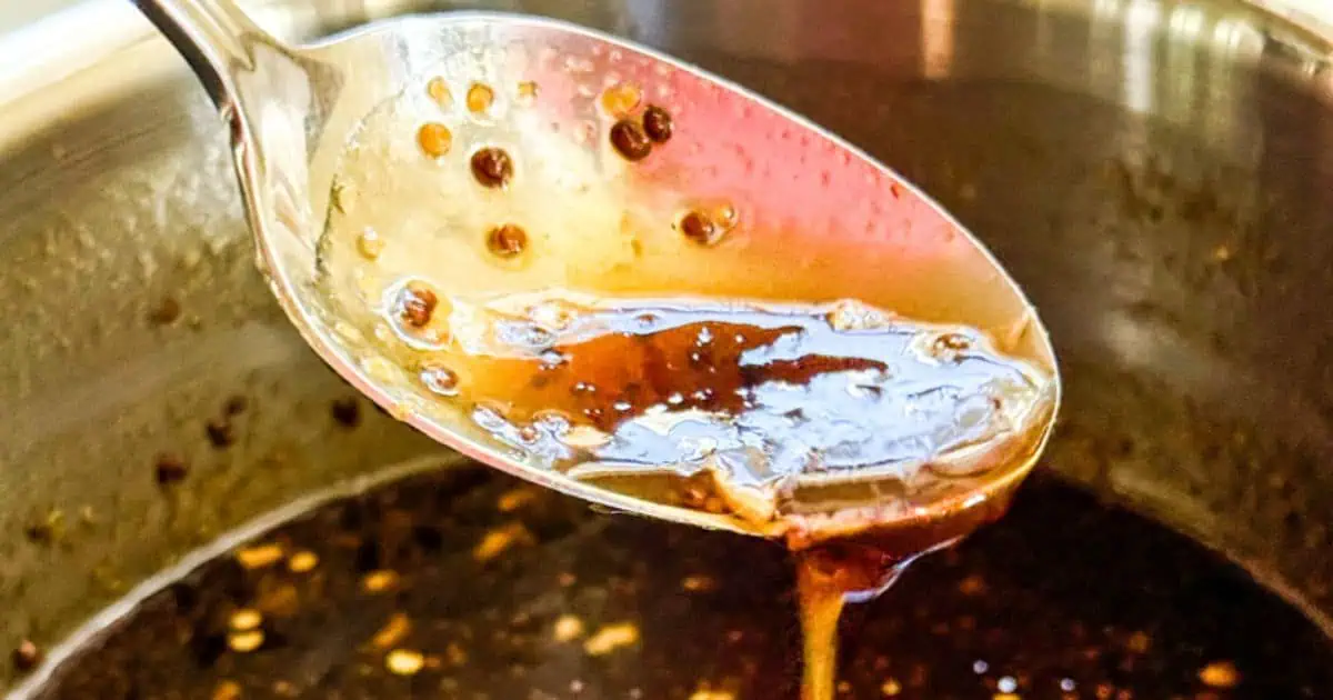 A close-up of a spoon dripping thick, dark soy sauce into a pot. The sauce appears to have small bits of seasoning or spices, adding texture to the liquid. The pot’s edges are partially visible in the background, indicating it's being used for cooking.