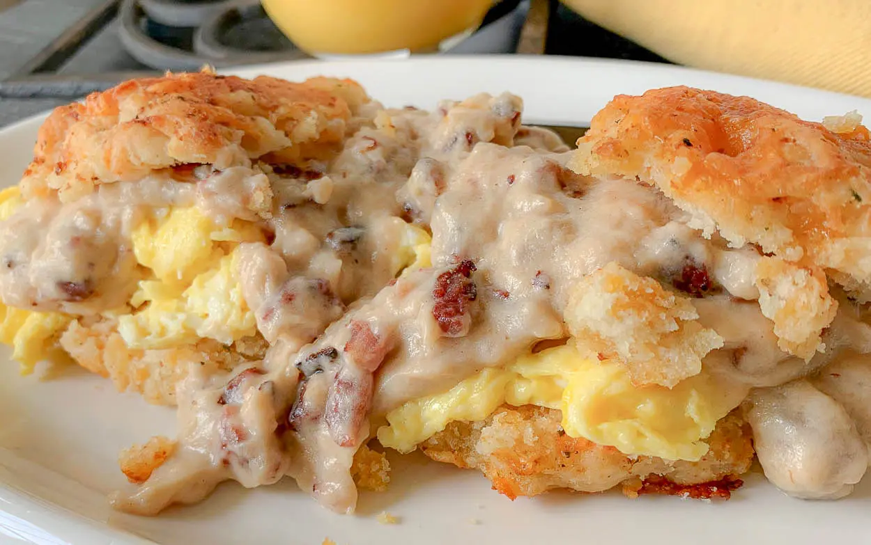 Two flaky cheesy biscuits are split open and filled with scrambled eggs, then topped with creamy sausage gravy containing visible bits of sausage, all served on a white plate—a classic Southern breakfast.