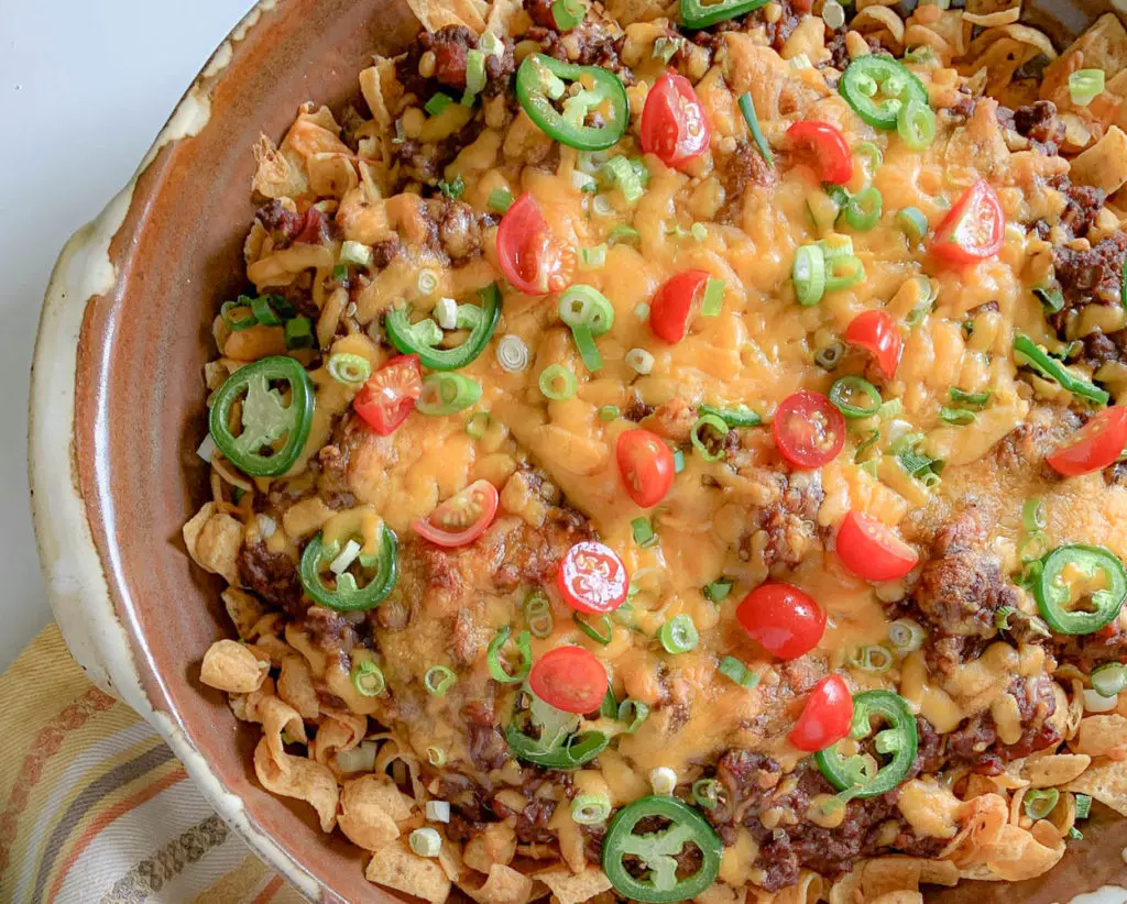 A dish featuring a hearty and colorful Frito pie. The casserole includes layers of corn chips, chili, melted cheese, cherry tomatoes, chopped green onions, and slices of fresh jalapeño, all baked in a rustic, round ceramic dish.