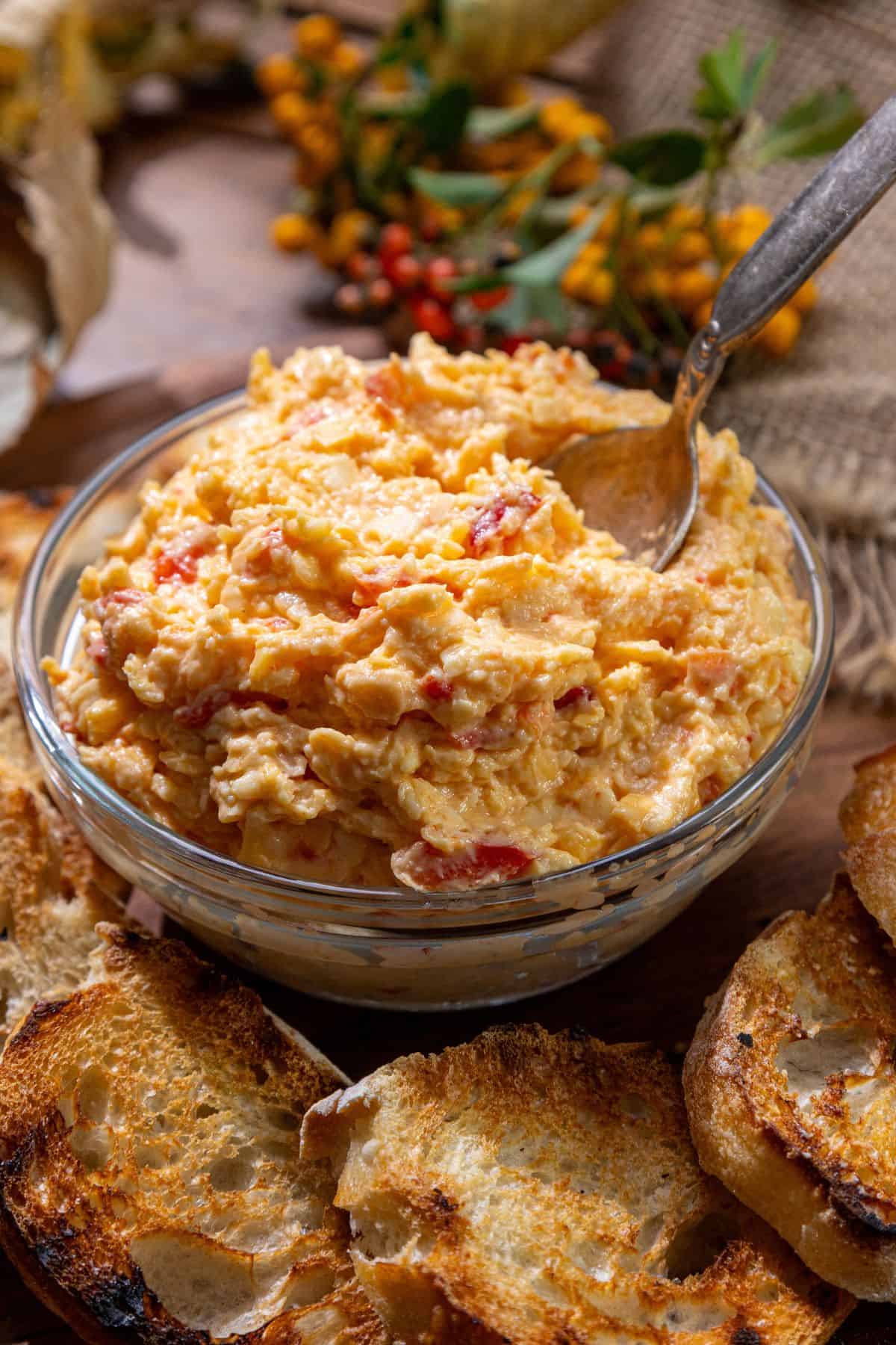 A glass bowl of white cheddar pimento cheese spread with a spoon, surrounded by toasted bread slices.