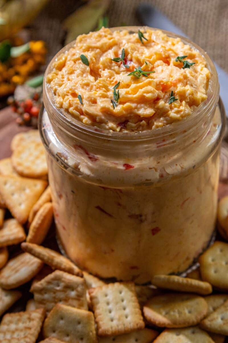 A glass jar of white cheddar pimento cheese spread sits on a wooden surface, surrounded by assorted crackers.