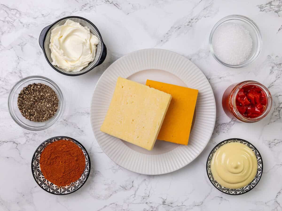 Two blocks of white cheddar on a plate, surrounded by bowls of cream cheese, mayonnaise, black pepper, paprika, salt, and roasted red peppers.