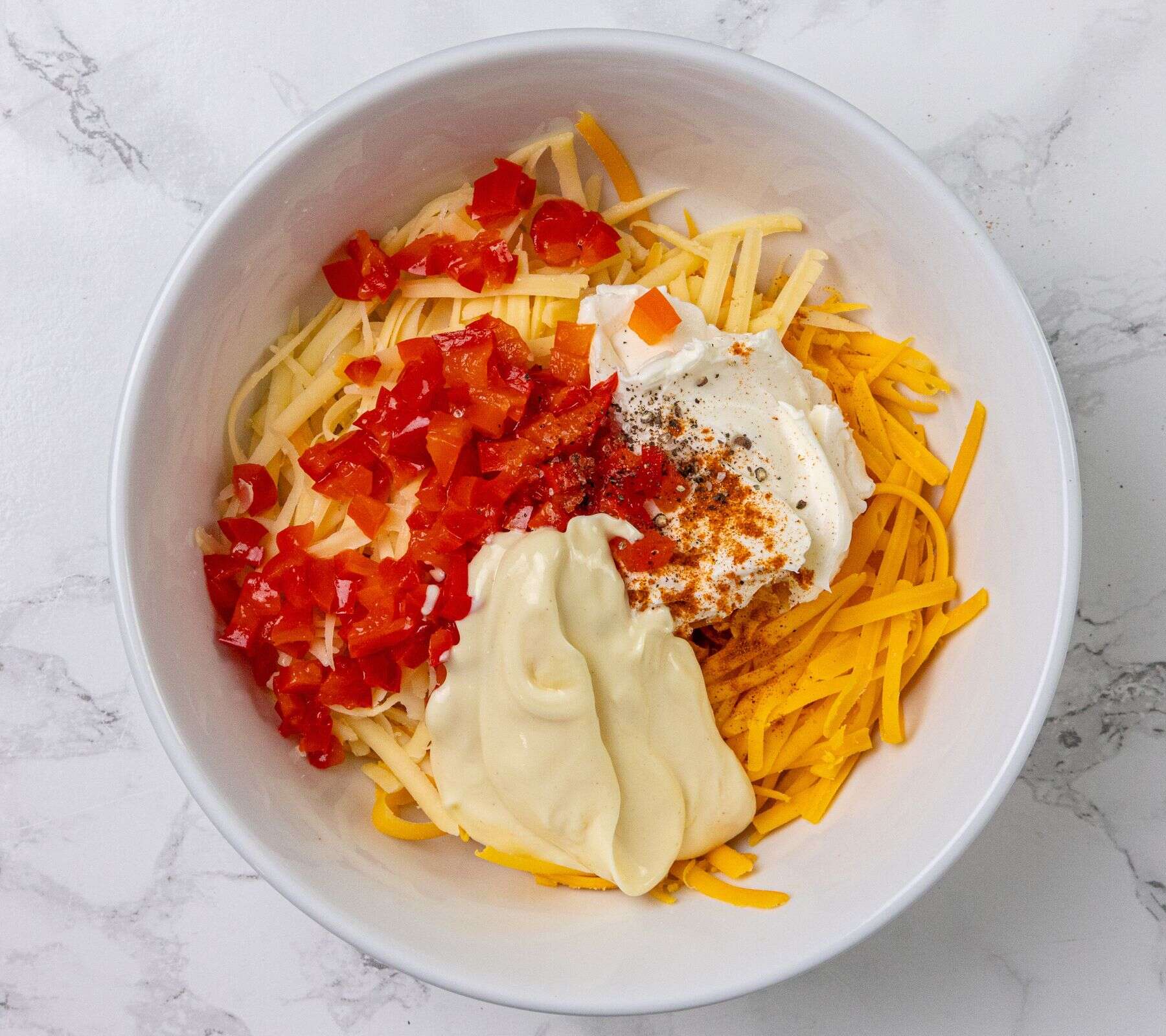 A white bowl of pimento cheese with visible chopped red peppers on a white marble surface.