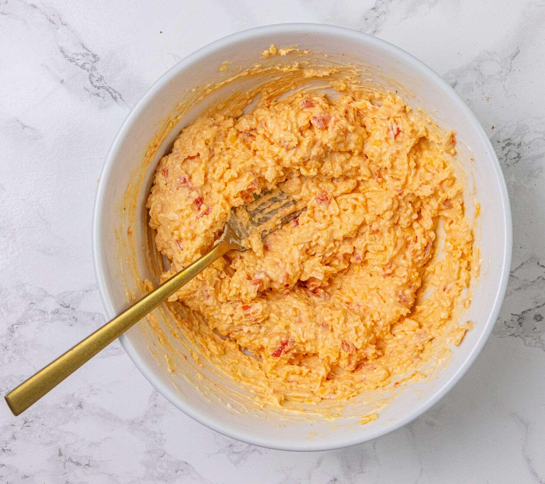 A mixing bowl with a gold fork and an orange, creamy pimento cheese mixture on a white marble surface.
