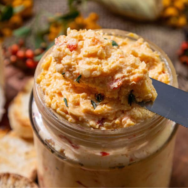 A close-up of a jar filled with white cheddar pimento cheese, with a knife scooping some out.