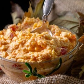 A glass bowl of white cheddar pimento cheese with a silver spoon rests on burlap, surrounded by autumn leaves.