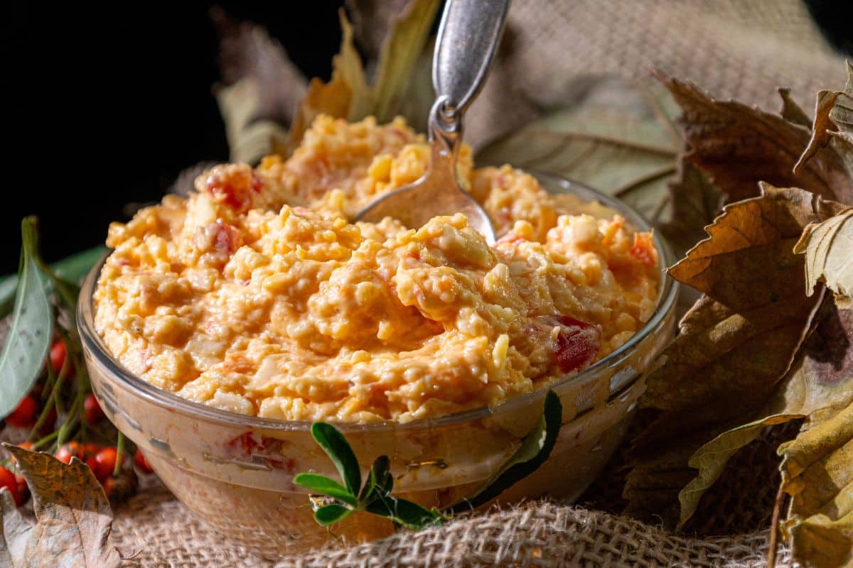 A glass bowl of white cheddar pimento cheese with a silver spoon rests on burlap, surrounded by autumn leaves.