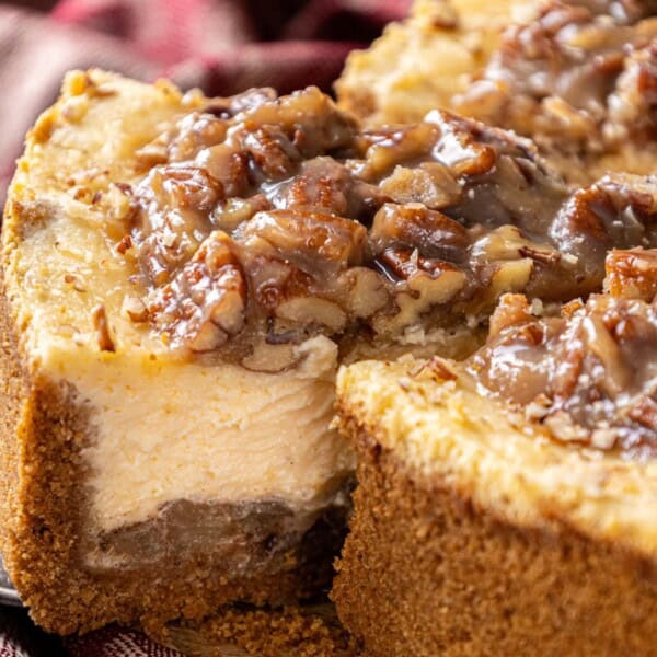 A cheesecake with a graham cracker crust, topped with pecans and sauce; a slice is being lifted from the whole cake.