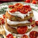 A stack of breaded eggplant rounds layered with white cheese and roasted tomatoes, garnished with herbs, on a patterned plate next to a gold fork and knife.
