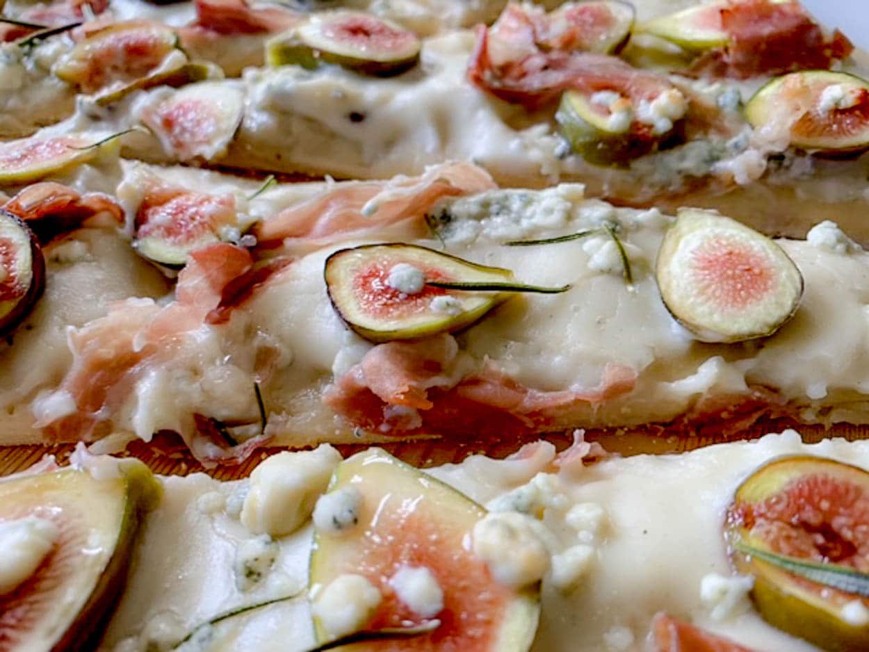 A close-up of a gourmet pizza topped with creamy cheese, slices of fig, prosciutto, and sprigs of fresh rosemary. The pizza has a rustic, artisanal appearance with a variety of textures and colors.