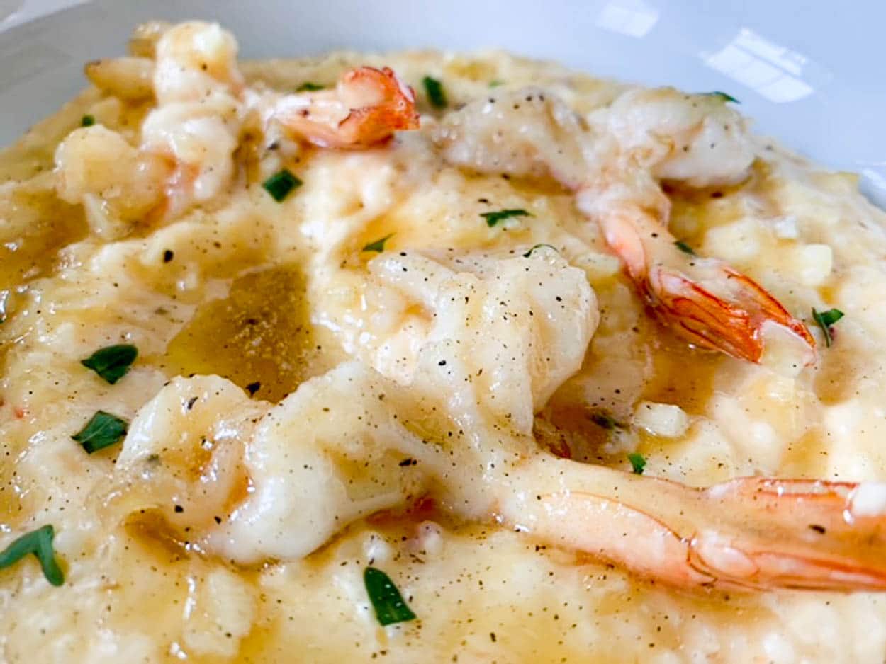 A close-up of a creamy shrimp and grits dish. The dish features large, sautéed shrimp laid on top of rich, buttery grits, garnished with green herbs and a drizzle of sauce. The grits appear smooth and slightly chunky, with a few pepper specks visible.