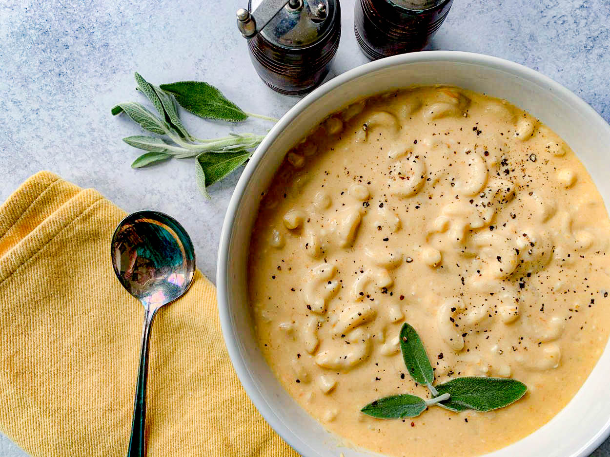 A bowl of creamy mac and cheese topped with black pepper and fresh sage leaves. A silver spoon rests on a yellow napkin next to the bowl, and two pepper mills are in the background on a light surface.