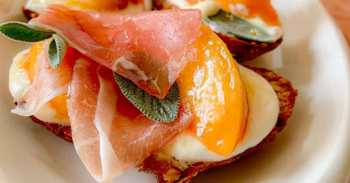 A close-up of appetizers featuring crispy bread topped with creamy cheese, slices of prosciutto, fresh peach slices, and sage leaves on a white plate.