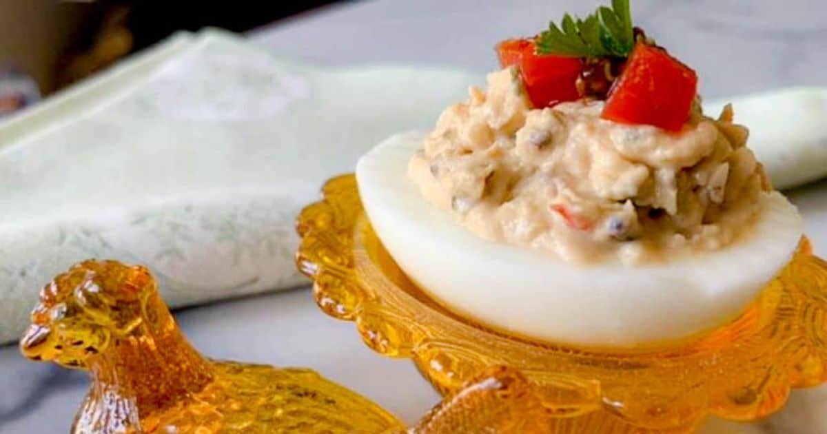 A halved boiled egg with creamy filling, topped with diced red peppers and parsley, rests on a decorative yellow glass dish shaped like a chicken. A folded white napkin is visible in the background.