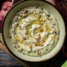 A bowl of whipped feta dip garnished with chopped herbs, nuts, and a drizzle of olive oil sits on a rustic wooden surface atop a red-patterned cloth.