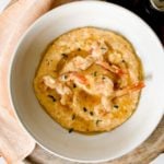 A bowl of shrimp and grits with chopped herb garnish on a wooden tray, beside a folded napkin and salt and pepper shakers.