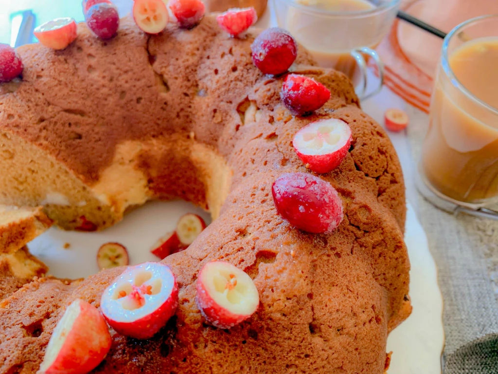 A festive bundt cake decorated with halved cranberries, displaying a golden-brown crust. Surrounding the cake are glasses of coffee and a light brown liquid, possibly eggnog or another drink. The cake sits on a white surface, creating a warm and inviting scene.