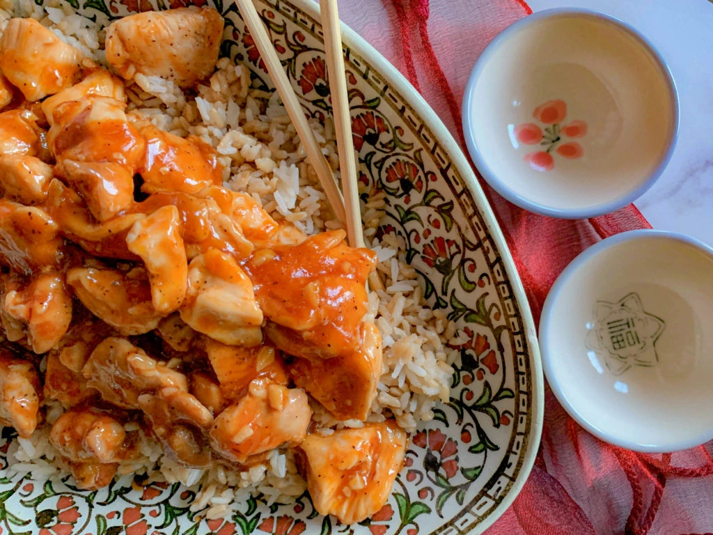 A decorative plate of rice topped with chicken in a spicy orange sauce, with chopsticks placed on the edge. Two small ceramic bowls with floral designs are situated on the side, along with a red cloth. The plate and chopsticks hint at an Asian-inspired meal.