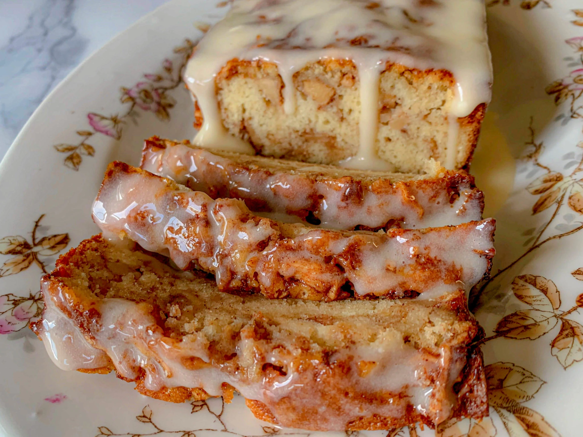 A delicious loaf cake covered in a glossy glaze sits on an ornate floral plate. Four thick slices have been cut from the loaf, showing a moist and crumbly texture with visible nut pieces and are drizzled with the same creamy glaze.