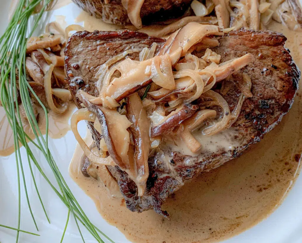 A thick, juicy steak topped with sautéed mushrooms and onions is drizzled with creamy sauce. Fresh chives garnish the dish, adding a touch of green. The steak appears perfectly cooked and the mushrooms and onions are tender, contributing to an appetizing presentation on a white plate.