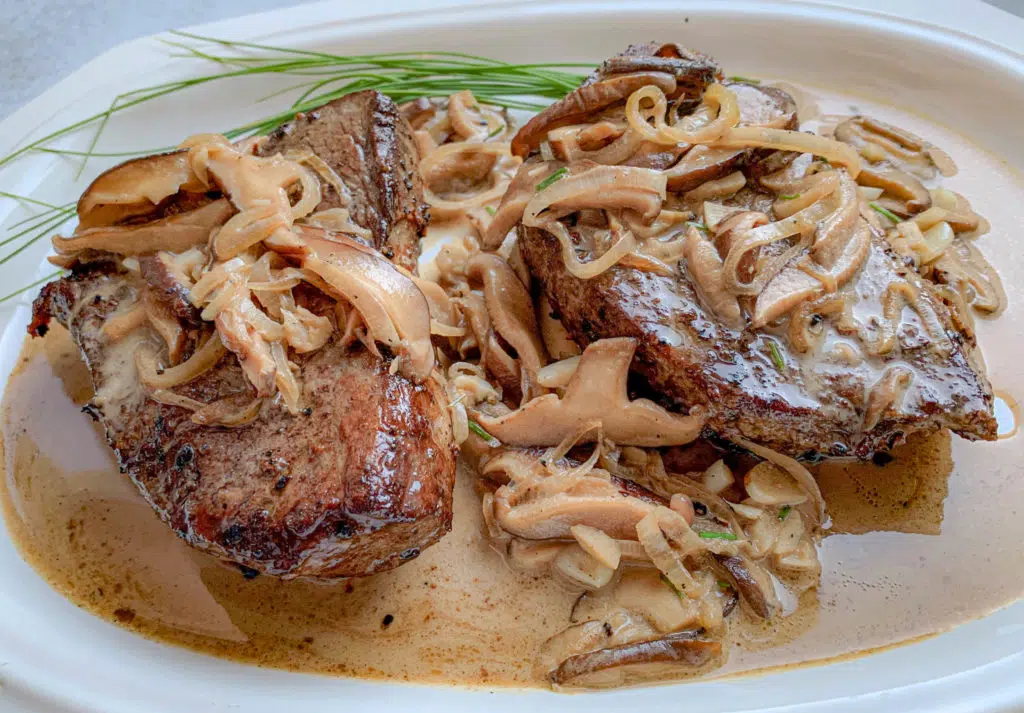 Two succulent pieces of beef topped with creamy sautéed mushrooms and onions, garnished with chives on a white platter.
