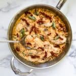 A skillet containing a creamy chicken dish with sundried tomato cream sauce, garnished with basil. A spoon is resting inside the skillet.