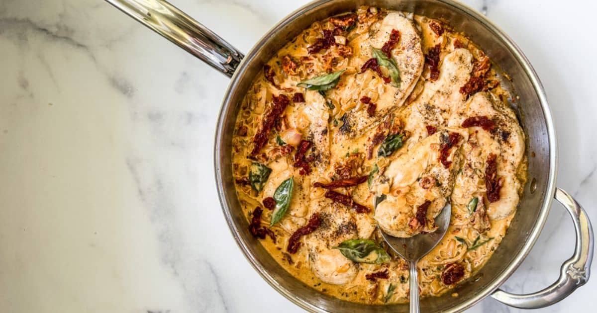 A skillet containing creamy chicken with fresh basil and a rich sundried tomato cream sauce, with a metal spoon resting in the dish.