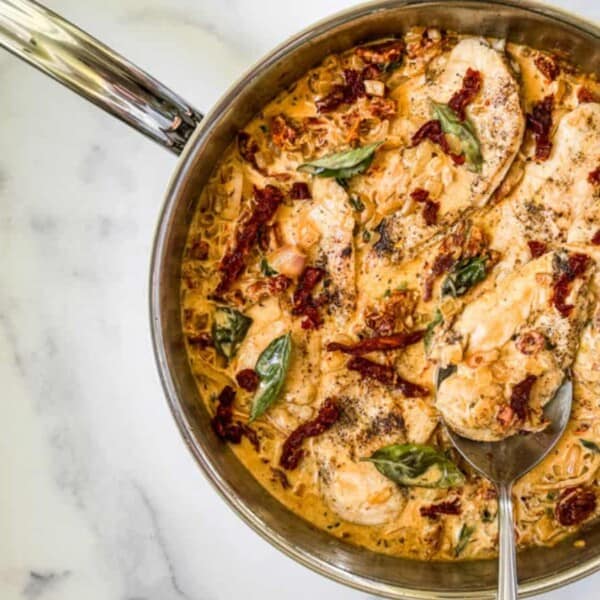 A metal pan with chicken and sun-dried tomatoes in a cream sauce, garnished with basil leaves, on a marble countertop.