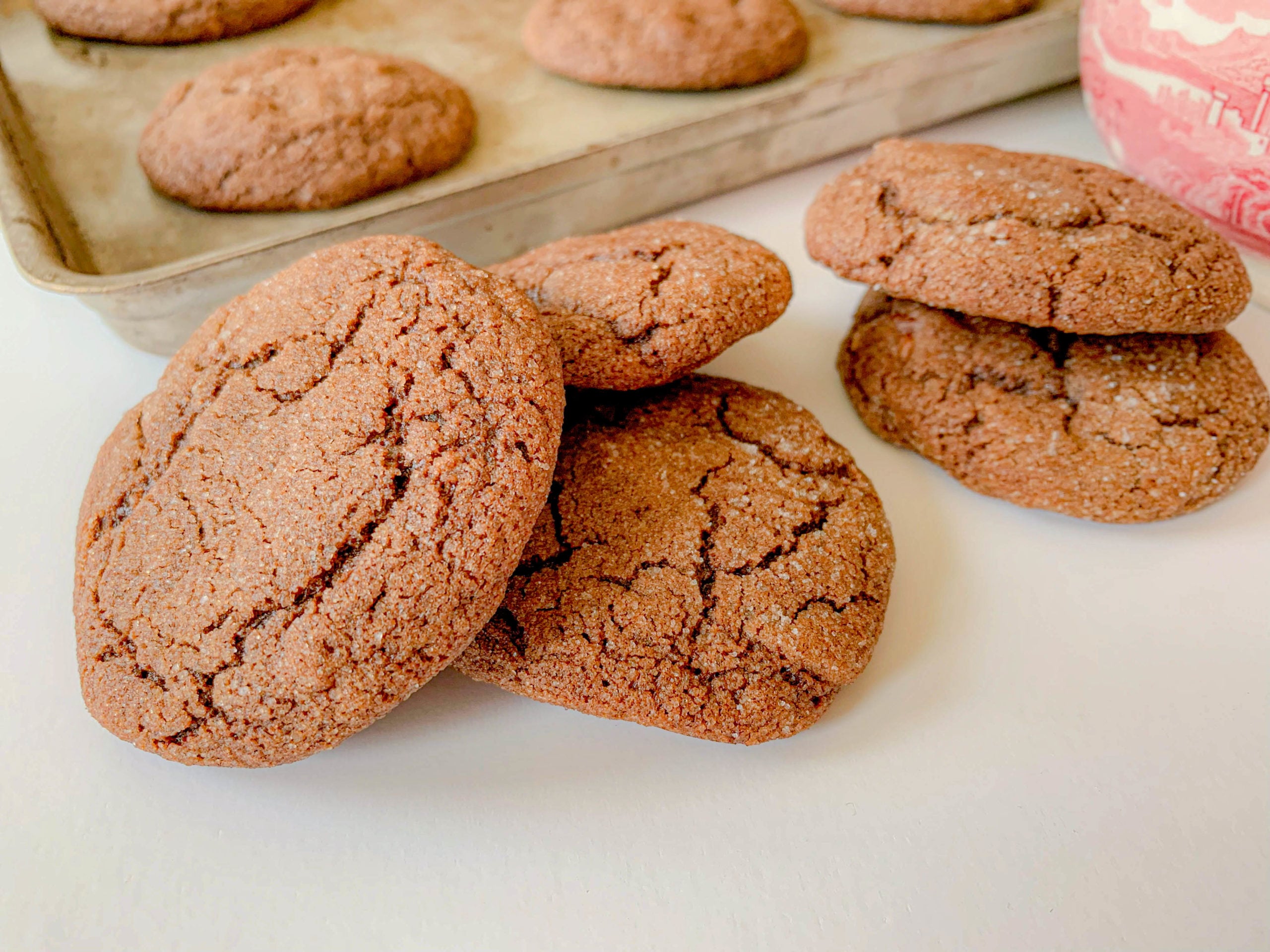 Close-up of freshly baked chocolate cookies on a white surface. A few cookies are stacked, while others are spread out. A baking tray with more cookies is visible in the background.