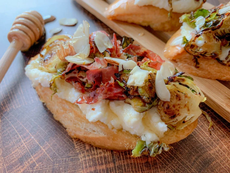 A slice of toasted baguette topped with creamy cheese, roasted vegetables, and sliced almonds, served with a honey dipper on the side. The wooden surface adds a rustic touch to the presentation.