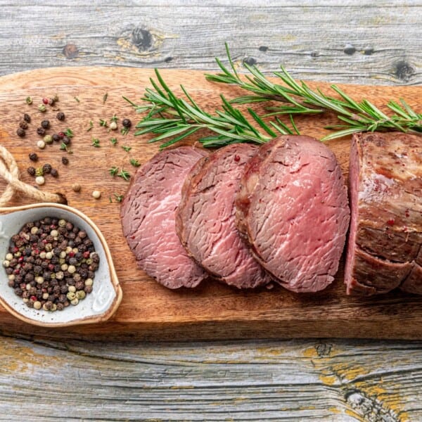 A sliced beef tenderloin roast with a rosemary garnish on a wooden board and a small bowl of mixed peppercorns beside it.