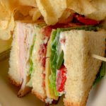 The image features layers of turkey, bacon, cheese, lettuce, and tomato between bread slices. Crispy potato chips are served alongside.