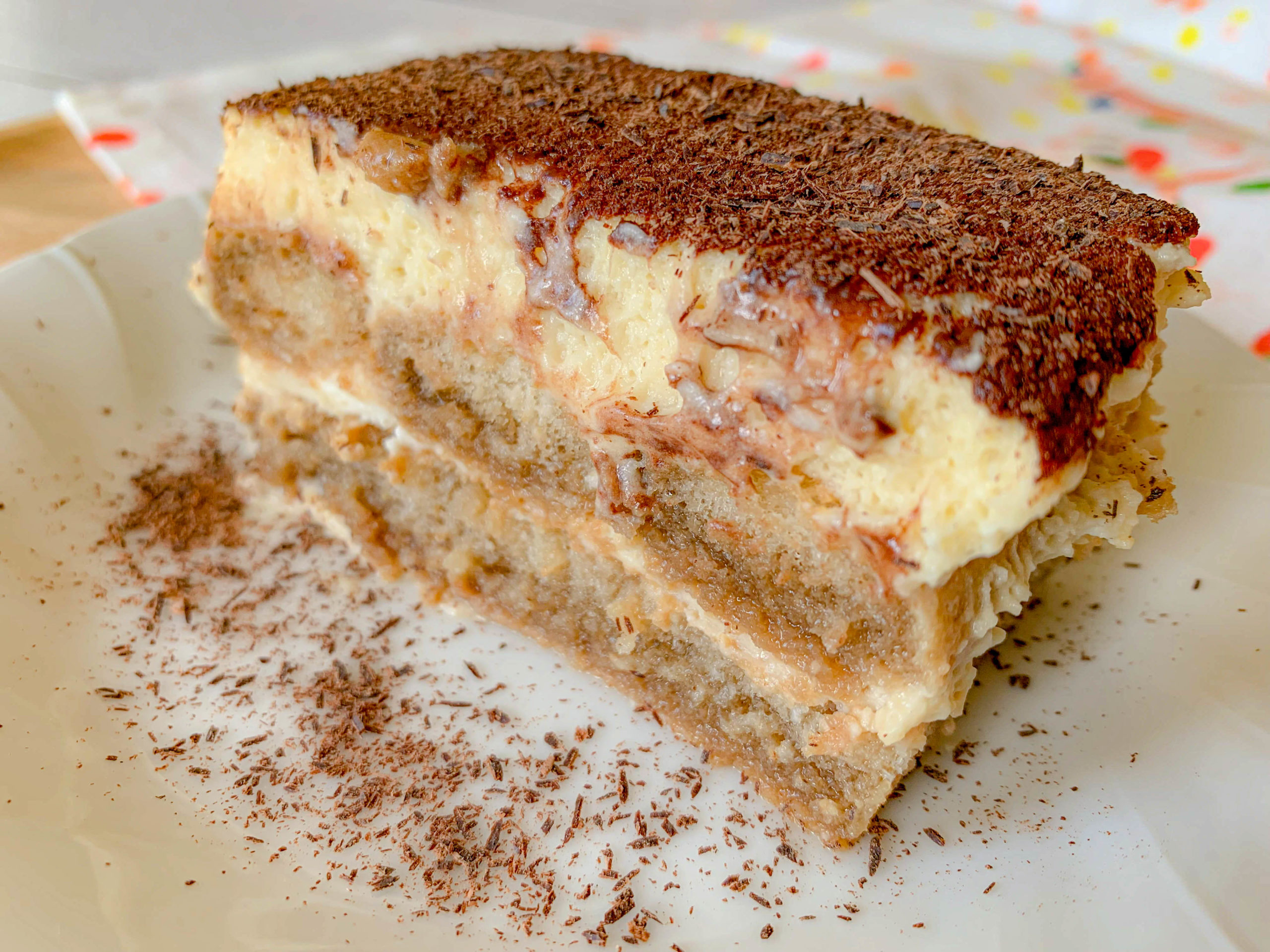 A slice of tiramisu on a white plate, topped with cocoa powder. Layers of espresso-soaked ladyfingers and creamy mascarpone are visible, with additional cocoa dusted around the plate.