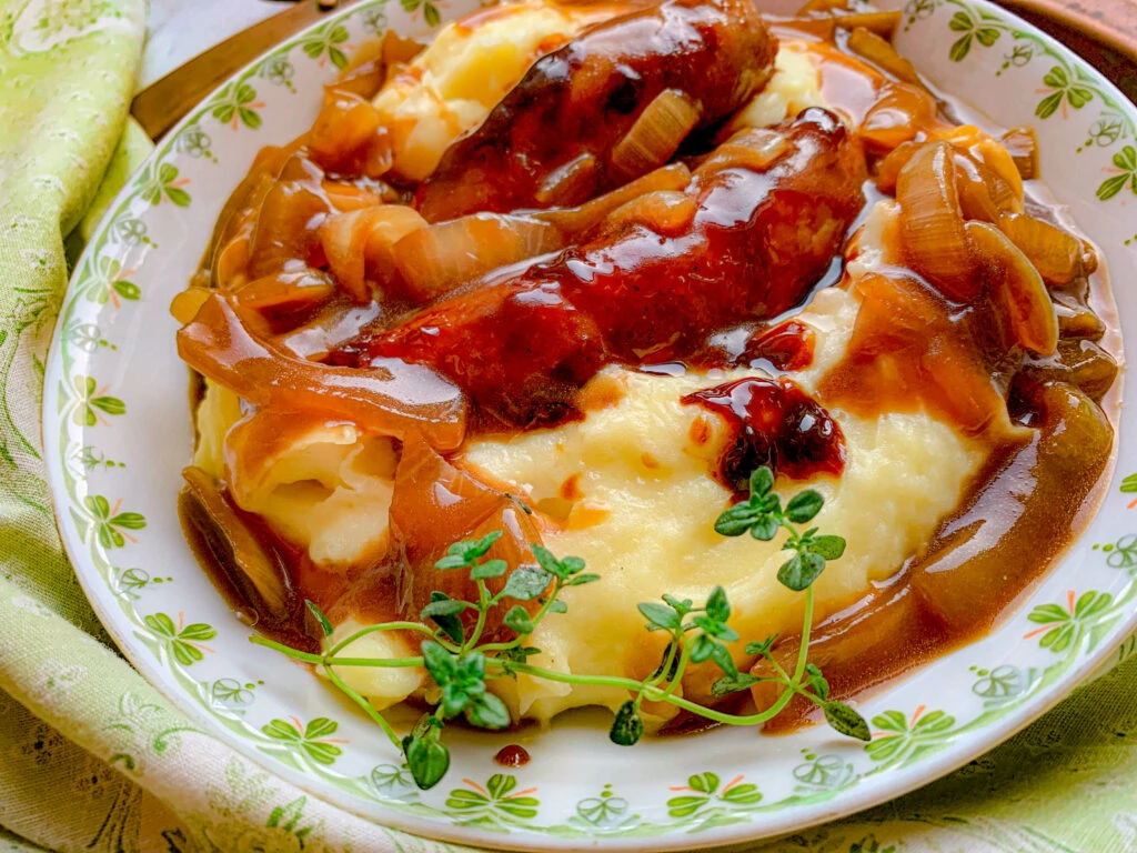 A dish of sausages and caramelized onions over mashed potatoes with gravy, garnished with fresh thyme, in a white bowl with a green floral border.