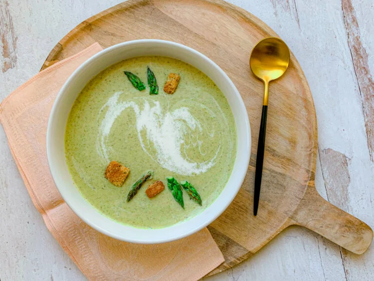 A bowl of creamy green soup garnished with asparagus tips and croutons, served on a round wooden board with a gold spoon. The board sits on a light surface next to a folded peach-colored napkin.
