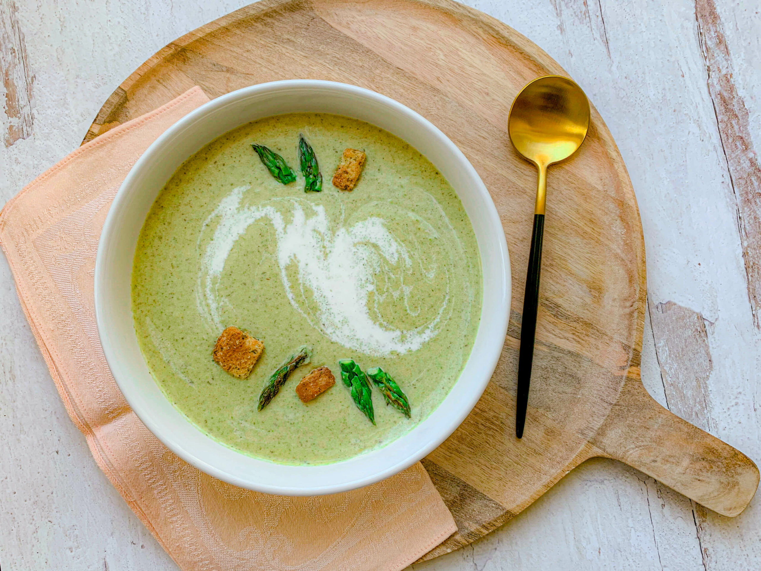 A bowl of green soup garnished with asparagus tips, croutons, and a swirl of cream, placed on a wooden serving board with a gold spoon and a peach napkin beside it. The background is a white rustic surface.