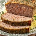 Sliced meatloaf topped with creamy onion gravy and herbs on a decorative plate, with a side of mashed potatoes.