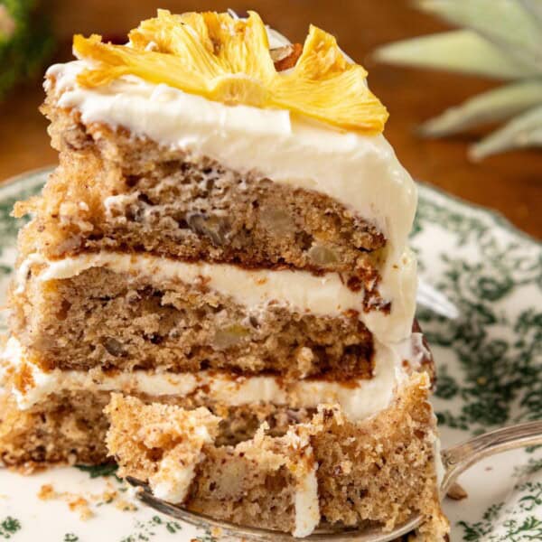 A slice of layered cake topped with frosting and dried pineapple sits on a decorative plate. A fork is nearby, surrounded by small flowers and pineapple leaves.