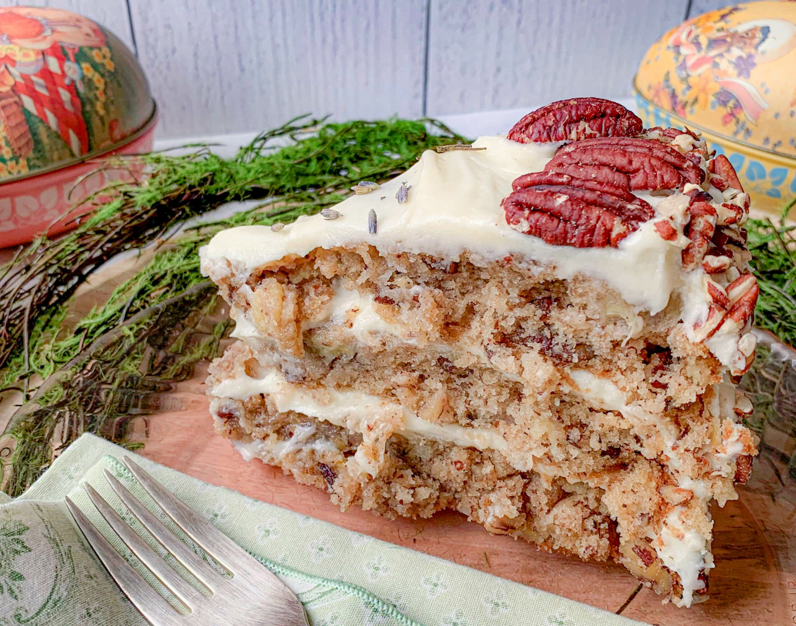 A generous slice of moist, frosted cake filled with nuts and topped with creamy icing and pecans is served on a wooden board. The cake is garnished with sprigs of greenery and accompanied by decorative ceramic containers in the background, alongside a silver fork on a patterned napkin.
