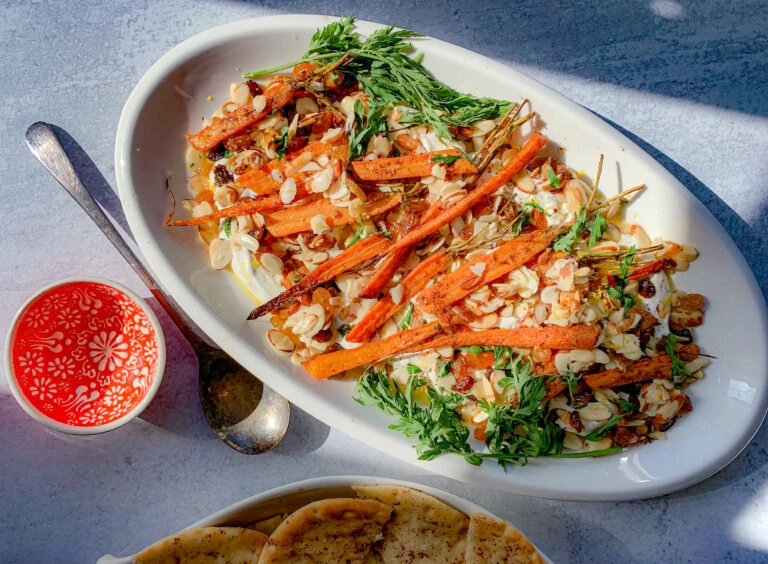 A vibrant dish featuring roasted carrots garnished with nuts and greens on a white oval plate. A red and white patterned cup of sauce and a spoon are beside it, accompanied by slices of naan bread on a light blue surface.