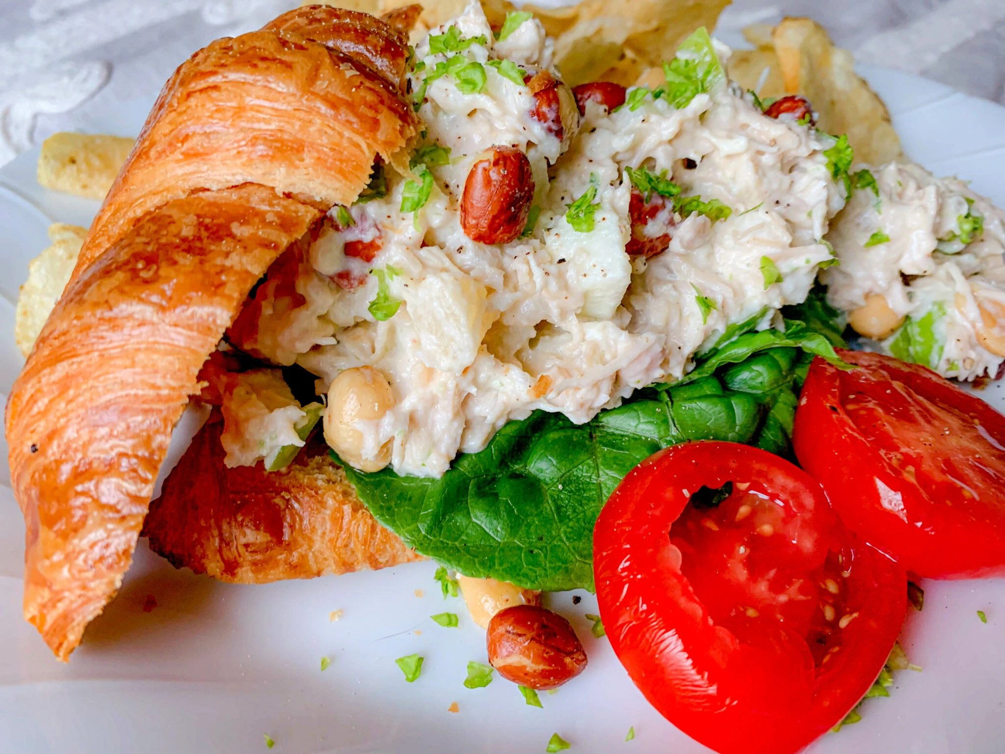 A croissant sandwich filled with chicken salad, featuring nuts and greens, is placed on a plate. The sandwich is accompanied by fresh spinach leaves on the same plate as slices of red tomato and a side of potato chips.