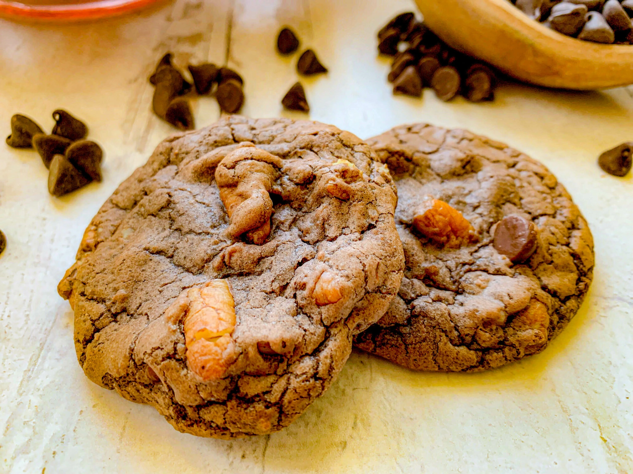 Close-up of two chocolate chip cookies on a light wooden surface. The cookies are slightly cracked and have a rich, chunky texture with visible chocolate chips. In the background, there's a bowl of chocolate chips and some scattered on the surface.