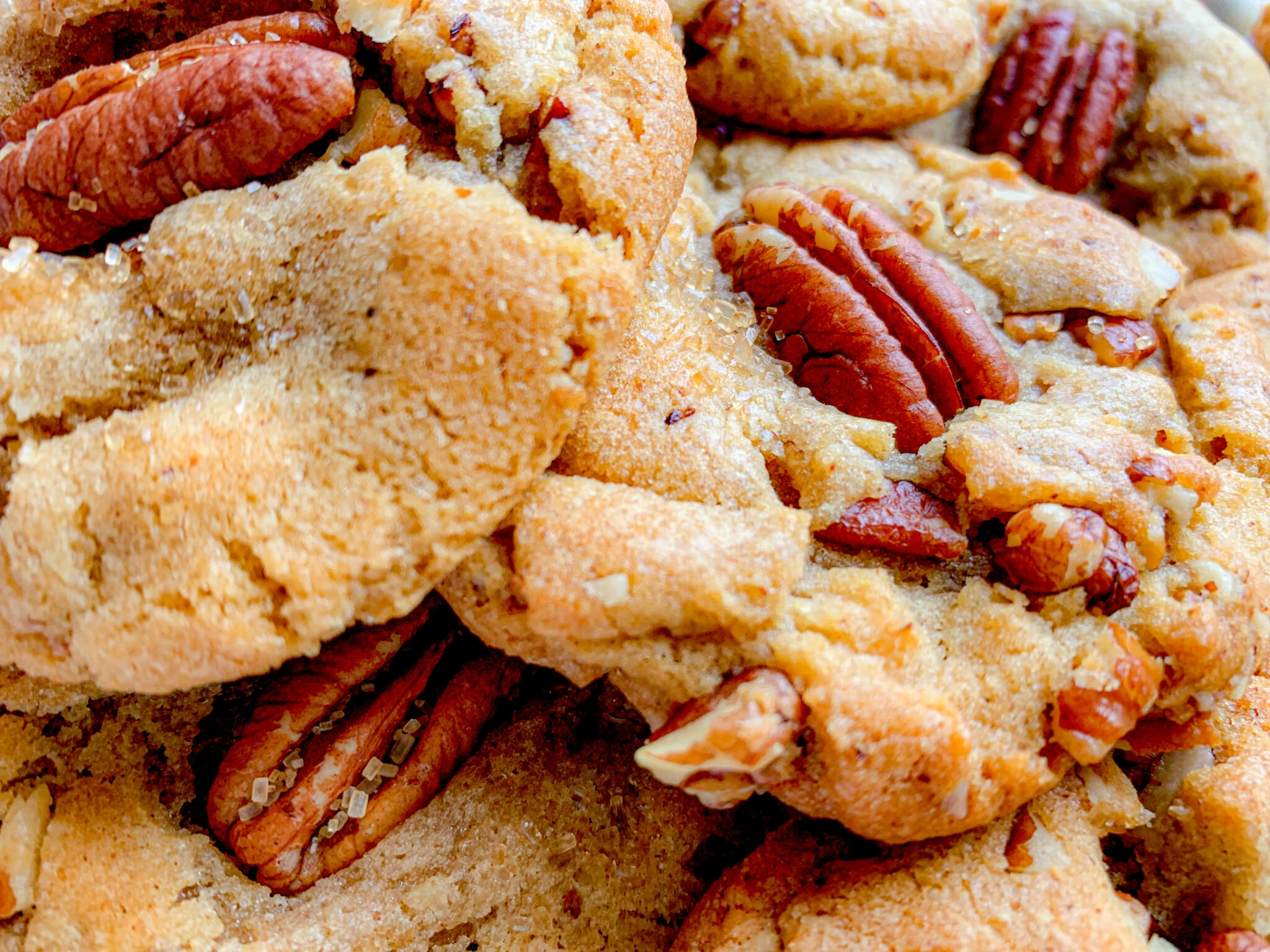 Close-up shot of a stack of freshly baked pecan cookies. The cookies are golden brown, with pieces of crunchy pecans visible on top and embedded within the dough. The texture appears soft and crumbly, showcasing a homely, homemade appeal.