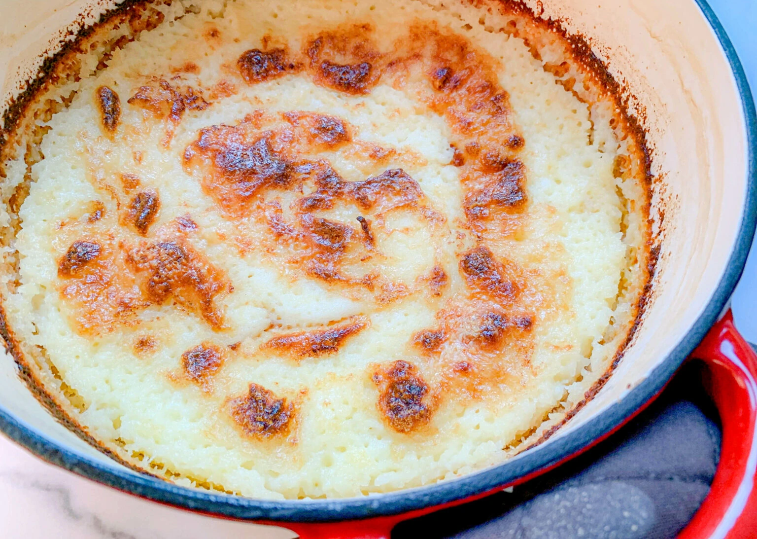 A close-up of a baked rice pudding in a round, red and white ceramic dish. The pudding has a golden-brown, slightly caramelized top with darker browned spots, indicating it has been oven-baked. The edges of the dish show some baked-on residue.