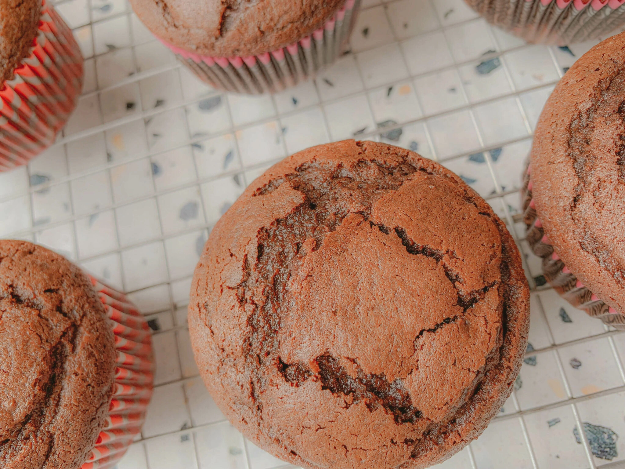 Close-up of several freshly baked chocolate muffins on a white, tiled surface. The muffins have a slightly cracked top and are in red and white striped paper liners.