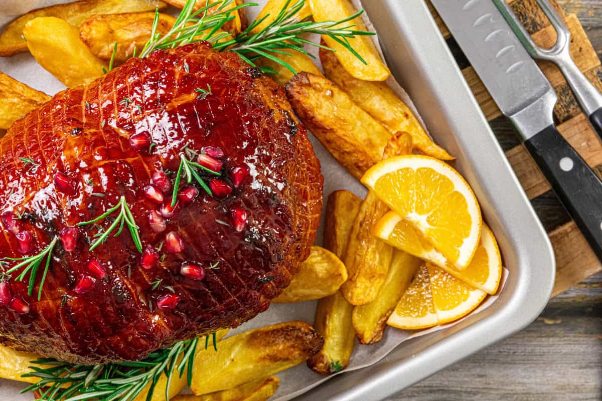 The image shows roasted ham topped with pomegranate seeds and rosemary. It is served with crispy potato wedges and fresh orange slices, all arranged on a baking tray. Nearby, knives are placed on a wooden chopping board.